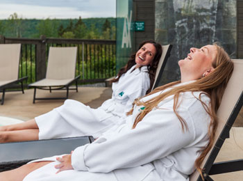 Two women in bathrobes laughing while lying on lounge chairs.