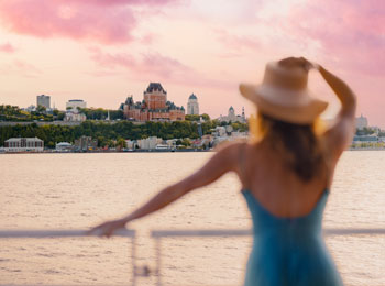 A woman in a blue dress and a large hat looks out over the Château Frontenac from the St. Lawrence River.