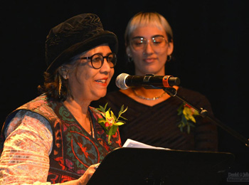 A woman wearing a hat recites a poem in front of a microphone and podium and a young woman stands beside her.