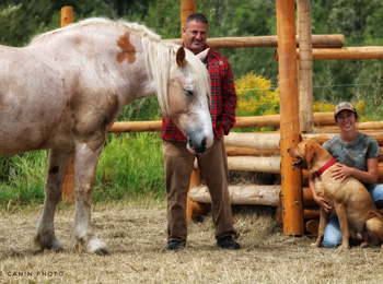 A man stands next to a horse, and next to him, a woman crouches with a dog.