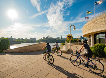 Mother and two sons on bikes next to the Canadian Museum of History in front of the Ottawa River.