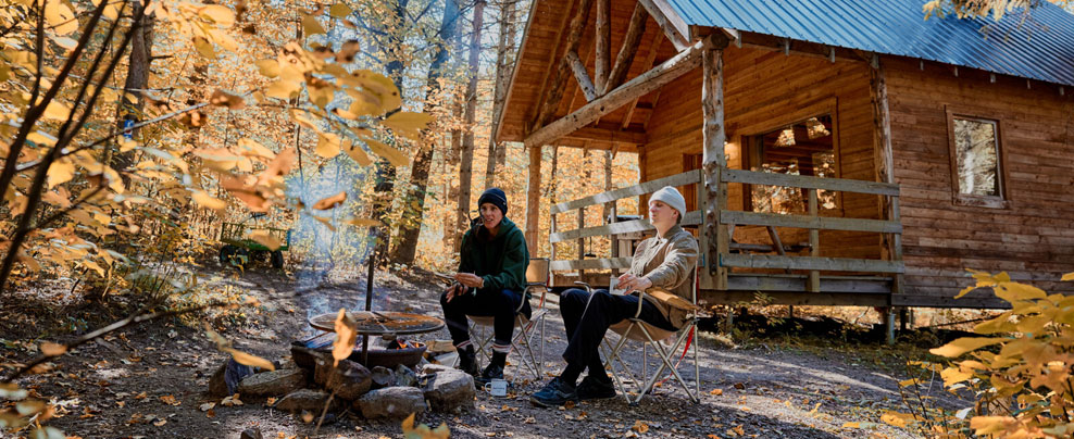 Two people sitting around a campfire in front of a wooden cottage in a forest in autumn.