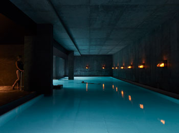 Indoor pool in a dark room lit only by soft lighting; a woman stands at the edge of the pool.