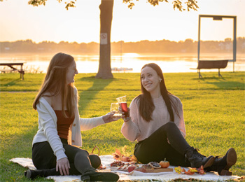 Two young women clink their glasses and share a picnic by the river,