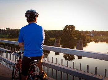 Man on bicycle seen from the back; he appears to be standing on a bridge over the water.